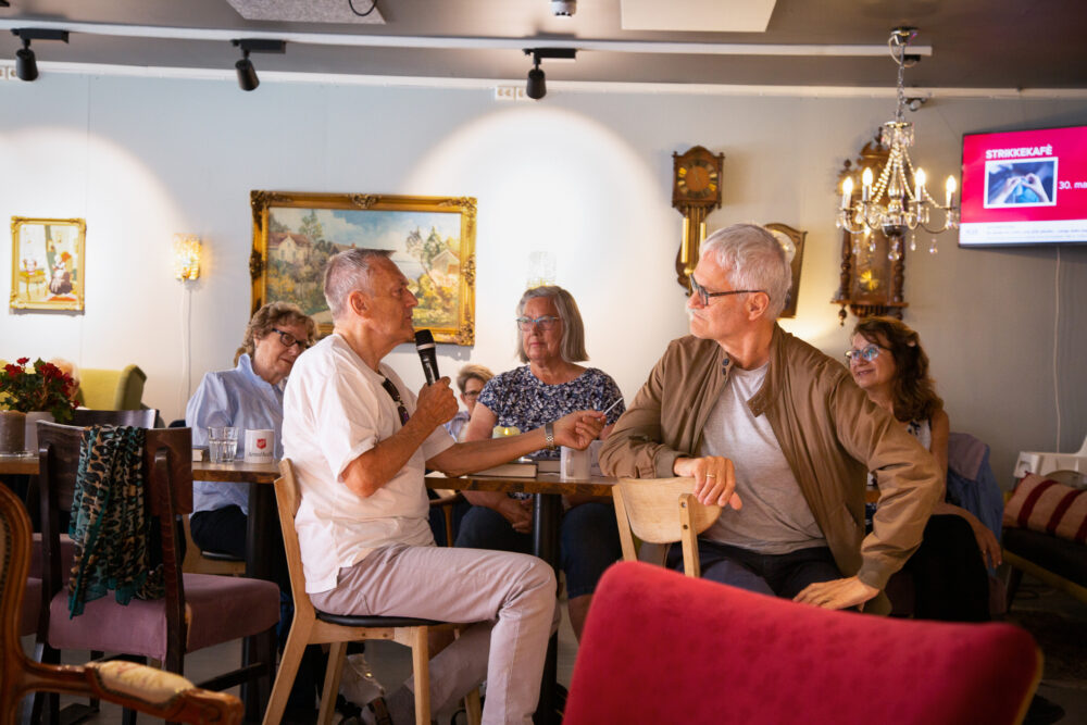 Gruppe med fem mennesker rundt kafebord. En mann i hvitt sitter og snakker i mikrofonen.