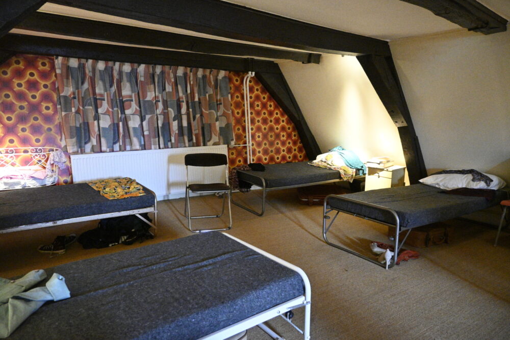 Et loftsrom med fire feltsenger langs veggene. Det er 70-tallstapet og -tekstiler på endeveggen og bjelker i taket