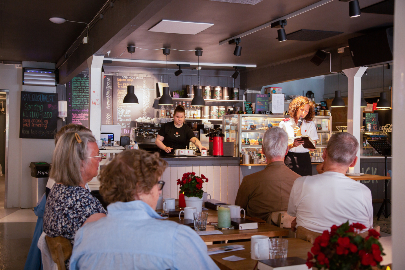 Folk ved mindre kafeborder ser fram mot en dame i uniform som snakker i mikrofon.