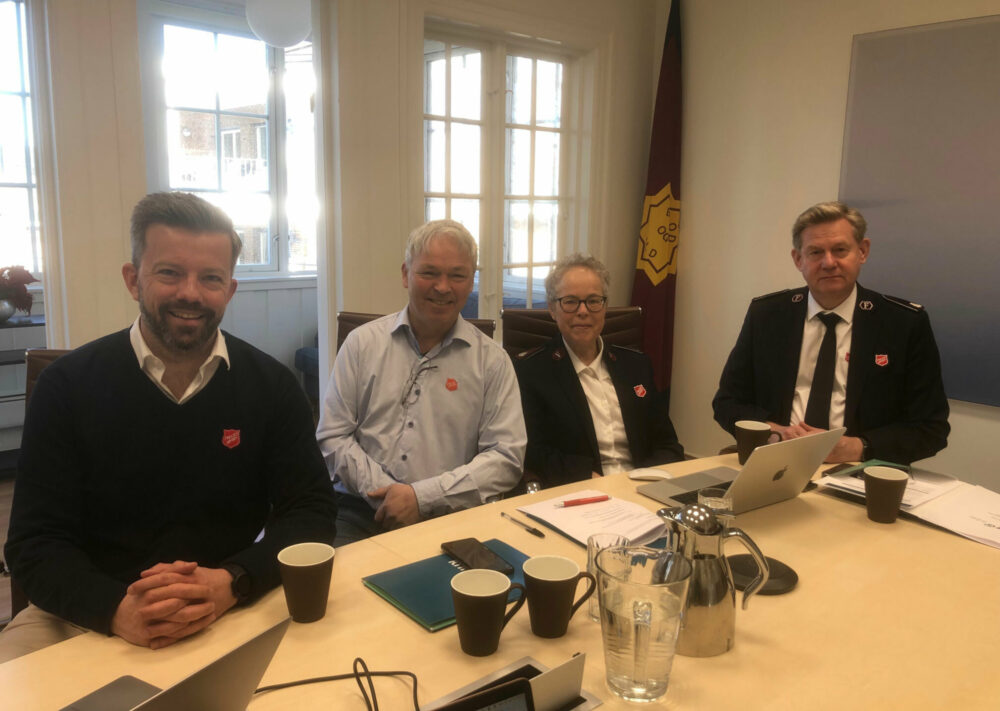 Foto: Tre menn og en kvinne sitter på rekke ved et bord. To av dem har på seg uniform fra Frelsesarmeen.