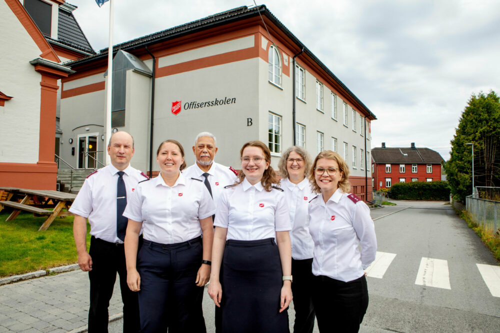 Foto: Utenfor et murbygg i beige og lakserødt står seks personer oppstilt. To menn og fire kvinner. Alle er kled i uniform fra Frelsesarmeen.