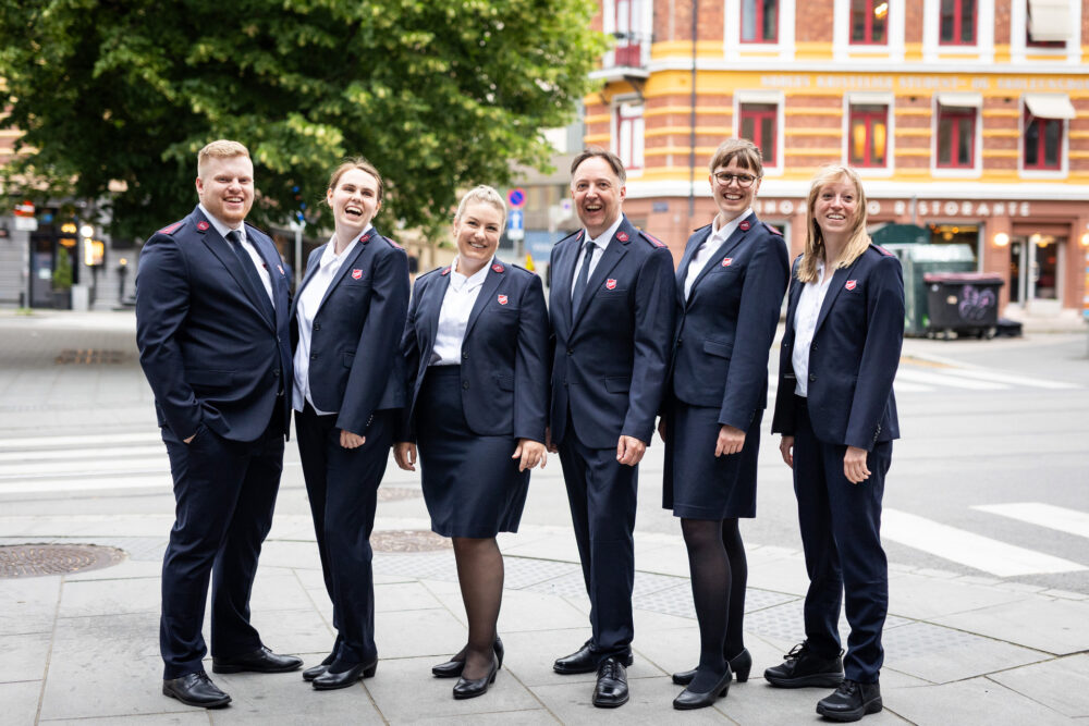 Foto: To menn og seks kvinner i uniformer fra Frelsesarmeen står på en rekke i bybildet.