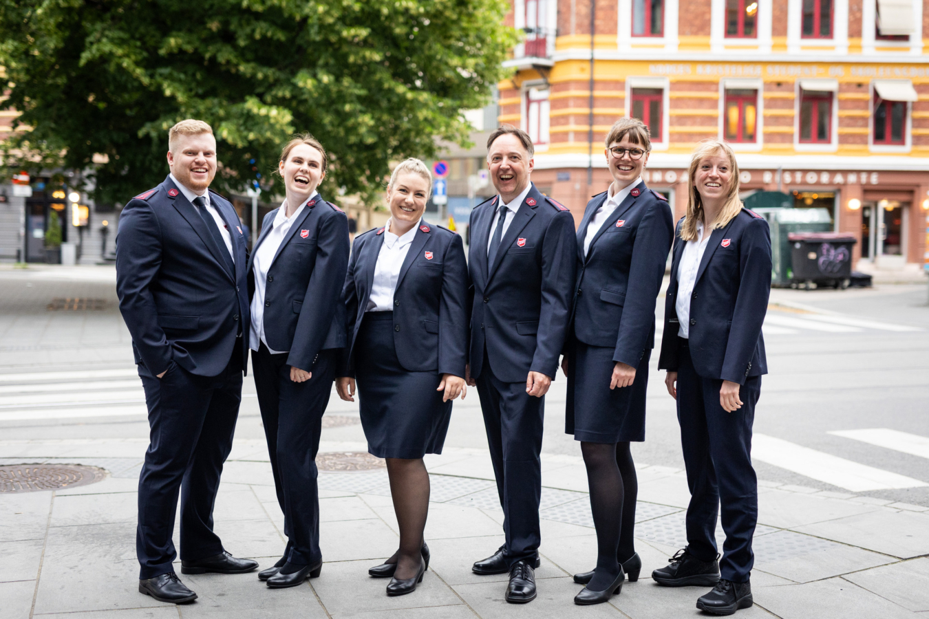 Foto: To menn og seks kvinner i uniformer fra Frelsesarmeen står på en rekke i bybildet.