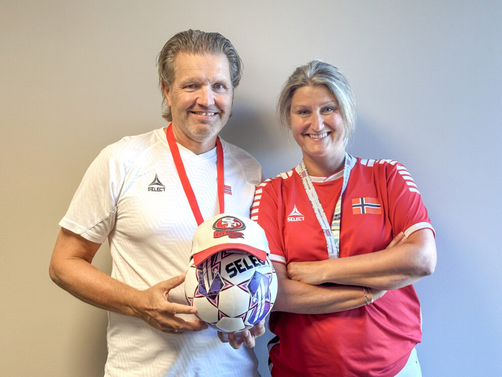 Foto: Mann i hvit trøye og kvinne i rød trøye står tett ved siden av hverandre og smiler. Han holder et fotball som har en caps på hodet. Begge har fløyter hengende i bånd rundt halsen.