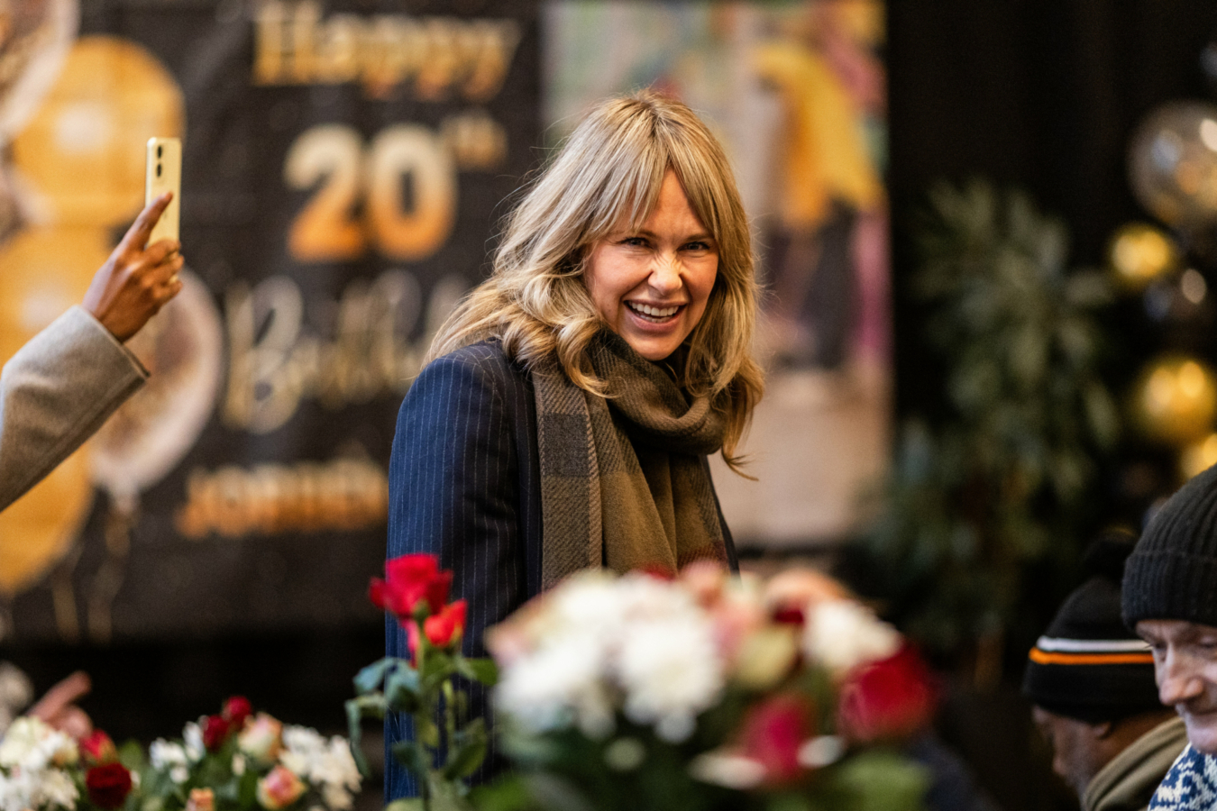 Foto: Blond kvinne i 50-årene med hår ned til skuldrene smiler til fotografen. På veggen bak henne henger en plakat der det står tallet 20. Foran er et bort med planter og julepynt.