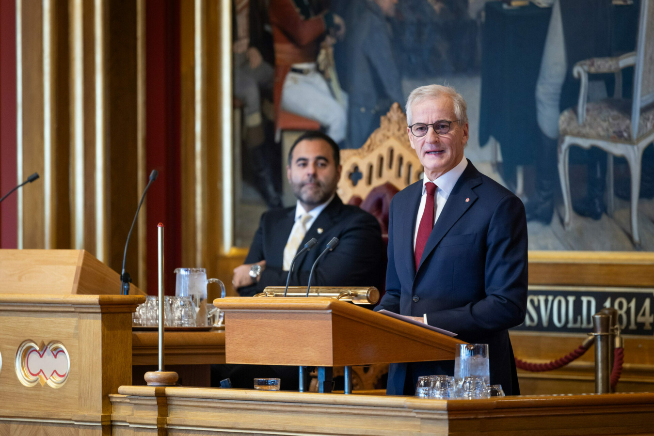 Foto: Statsministeren står på talerstolen i blå dress. Han har på seg lesebriller der han står på talerstolen i Stortinget. Litt bak ham sitter stortingspresidenten. Han har på seg svart dress med lysegult slips.